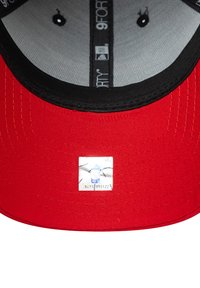 Gorra de béisbol roja con visera negra. Presenta un forro interior gris, etiquetas de logo y una pequeña etiqueta de autenticidad en la parte inferior de la visera.