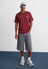 T-shirt a maniche corte color bordeaux con logo, pantaloni corti in denim grigi, calze bianche alla caviglia e sneakers multicolori. Gli accessori includono un cappellino a righe e occhiali da sole.