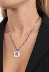 Collana d'argento con pendente circolare con accenti incisi, attaccata a una catena con anelli, indossata sopra un indumento nero.