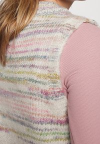 Gilet lavorato a maglia color avorio con strisce multicolori, caratterizzato da un tessuto testurizzato e un collo alto. Indossato sopra una maglietta rosa chiaro.