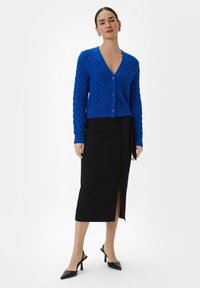 Blauwe gebreide cardigan met een gegolfd zigzagpatroon, gecombineerd met een zwarte midi-skirt met een zijsplit, afgemaakt met zwarte puntige hakken.
