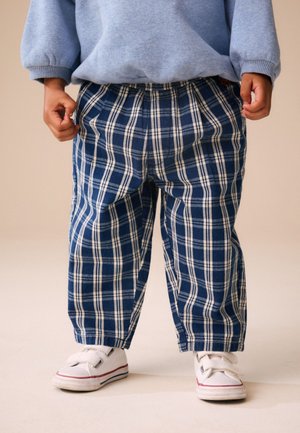 Barn iført blå og hvide ternede bukser, hvide sneakers med velcrolukning og en lyseblå sweatshirt, står på en neutral baggrund.