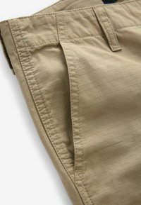 Beige bukser lavet af et let, struktureret stof med et rutenetmønster. Inkluderer standard lomme-design og synlige syninger som dekorative detaljer.