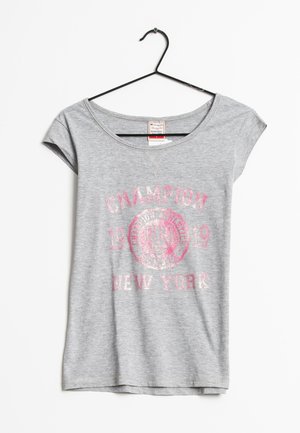 Lysegrå T-shirt med korte ærmer med lyserød falmet tekst "CHAMPION 1919 NEW YORK" og logo, hængende på en sort bøjle mod en hvid baggrund.