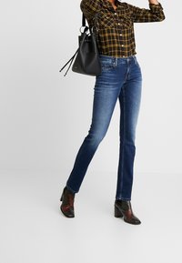 Blauwe denim jeans met een slim fit, gecombineerd met een zwarte bucketbag. Het model draagt een geruit shirt en enkellaarsjes met hakken met rode accenten.