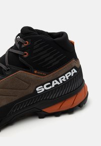 Pārgājienu zābaki ar melnu un brūnu krāsu shēmu, ar GORE-TEX etiķeti, teksturētu virspusi, mīkstinātu apkaklīti un dzīvīgu oranžu akcentu.