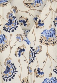 Florale Bluse mit weißem Stoffhintergrund, versehen mit blauen und beigen Blumenmustern und einem tiefen V-Ausschnitt. Weiche Textur mit leichtem Fall.