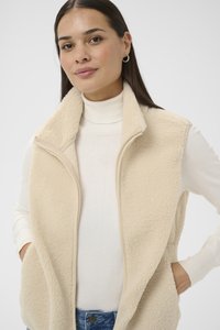 Femme portant un gilet sherpa crème par-dessus un pull à col roulé blanc, les mains dans les poches du gilet, debout devant un fond clair uni.