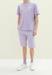 Camiseta de algodón de color púrpura claro con mangas cortas y un gráfico frontal, combinada con pantalones cortos a juego y zapatillas blancas. Diseño casual de color sólido.