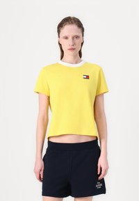 Gul bomulds T-shirt med hvidt rund halsudskæring, korte ærmer og et lille flerfarvet logo-patch på brystet, parret med mørke shorts.