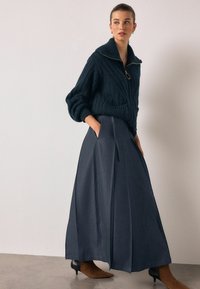 Tumesinine, kootud kampsun kõrge krae ja tõmblukuga, määratud halli, pika voldilise seelikuga. Komplekti lõpetavad pruunid terava ninaga kontsakingad.