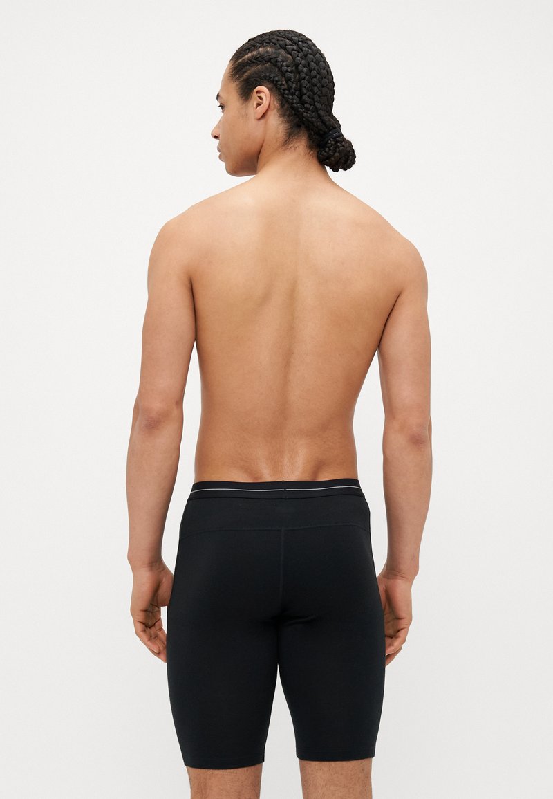 Pantalones cortos de tela elástica negra con un ajuste ceñido y cintura elástica, con acentos en gris. Usados por un modelo que muestra la vista trasera.