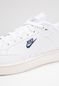 Zapatilla de cuero blanca con logo de Nike bordado en navy, cordones texturizados blancos y suela de goma color crema. Presenta detalles de perforación en los laterales.
