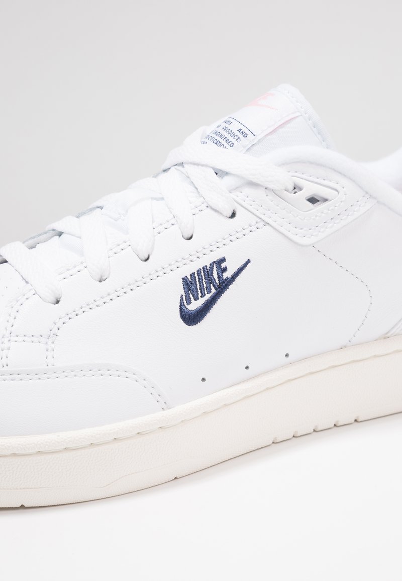 Sneaker en cuir blanc avec logo Nike brodé bleu marine, lacets texturés blancs et semelle en caoutchouc crème. Présente des perforations sur les côtés.