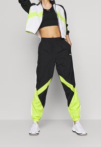 Woman in black sports bra, white and neon yellow black track jacket, matching pants, and white sneakers standing against a plain background.