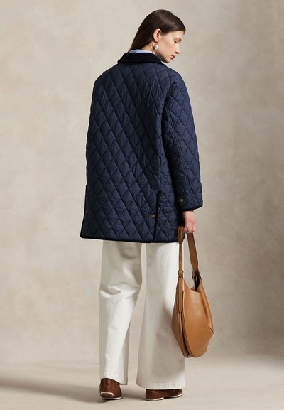 Femeie purtând o geacă matlasată bleumarin și pantaloni albi, ținând o geantă de umăr din piele maro deschis, stând în picioare pe un fundal neutru.