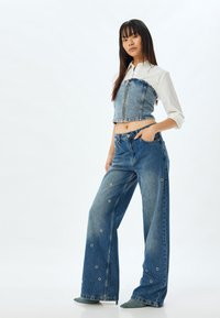 Top corto de mezclilla con cremallera frontal y jeans de pierna ancha con detalles de ojetes circulares. Ambas prendas en un lavado azul medio con un acabado mate.