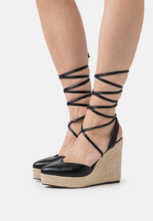Black leather wedge espadrilles feature a woven jute heel, open back, and long black laces wrapped around the ankles. Smooth texture throughout.