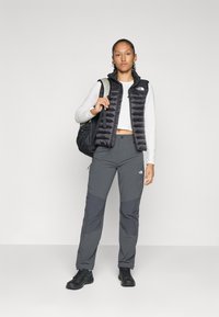 Svart puffy vest, grå langermet skjorte, mørkegrå bukser med lysere detaljer, svart ryggsekk og svart fottøy, stående på en enkel bakgrunn.