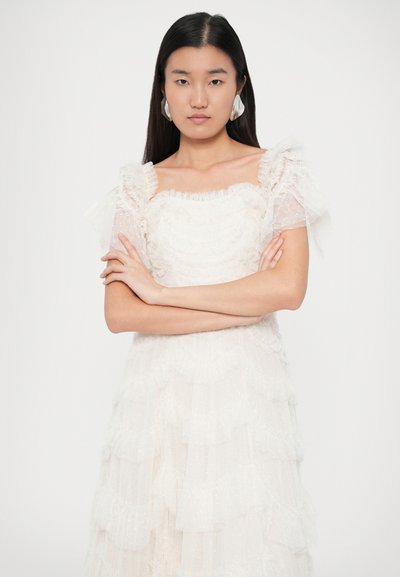 Bílé vrstvené šaty s volánovými rukávy, vyrobené z texturovaného materiálu a s měkkými, průhlednými detaily. Modelka má na sobě velké stříbrné náušnice.