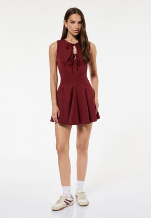 Femme portant une robe bordeaux sans manches avec des liens à l'avant, des chaussettes blanches et des baskets beiges, debout devant un fond blanc uni.