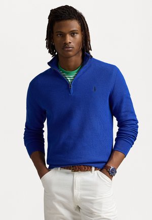 Pull-over bleu à col zippé avec des poignets côtelés, en tissu texturé, et accent logo. Porté sur une chemise rayée verte, associé à un pantalon blanc.