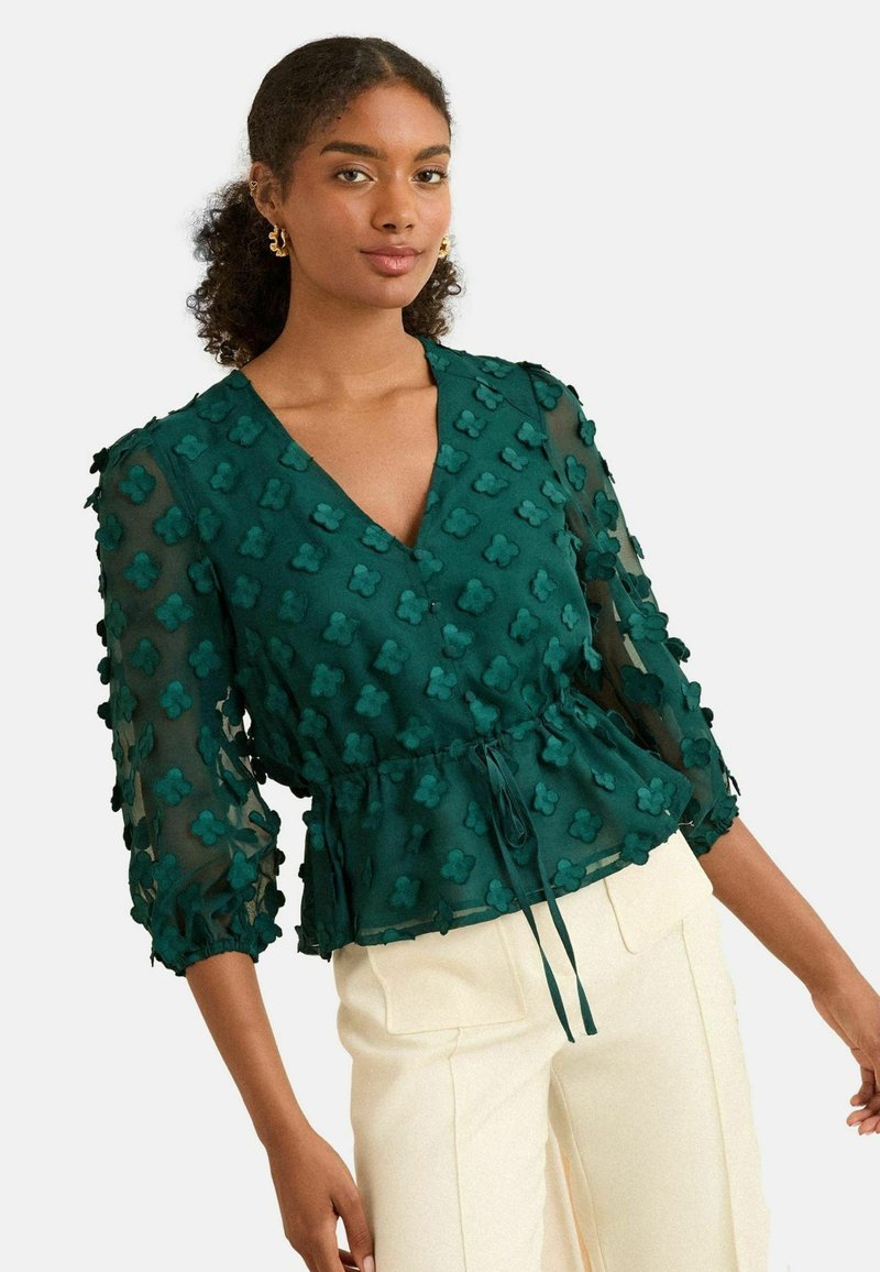 Blusa teal in tessuto trasparente, maniche a 3/4, motivo floreale applicato, scollatura a V, vita arricciata con dettaglio a fiocco. Abbinata a pantaloni di colore chiaro.