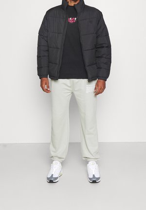 Svart vadderad jacka, svart T-shirt med grafik, ljusbeige mjukisbyxor med vit text och grå/vita sneakers. Står på en neutral bakgrund.