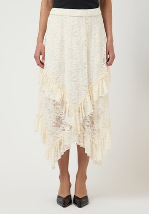 Kvinne som har på seg et kremfarget midiskjørt i blonder med rysjekanter og asymmetrisk hem, kombinert med svarte, spisse hæler.