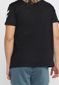 Sort kortærmet T-shirt med løs pasform, som har en glat tekstur og hvide chevron-detaljer på venstre skulder.