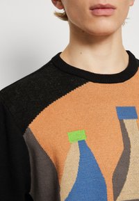 Närbild av en person som bär en svart, orange och grå tröja med abstrakta stickade flaskformer i blå, beige, grön och grå färg.