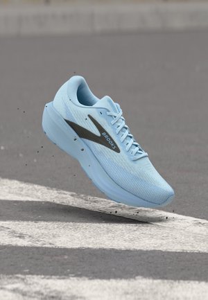 Scarpa da corsa Brooks azzurro chiaro con logo nero, lacci e suola spessa sospesa sopra una strisce pedonali su una superficie di asfalto grigio.