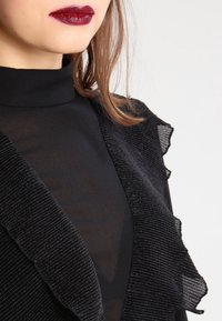 Top nero con collo alto e bordi arricciati, realizzato in materiale trasparente con un design testurizzato, che mette in risalto una leggera lucentezza.