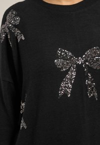 Schwarzer Pullover mit silbernen Paillettenbogen-Designs auf den Schultern. Weiche Textur, runder Ausschnitt und lockere Passform.