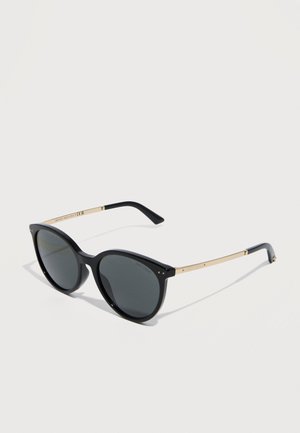 Črna okrogla sončna očala z rjavimi lečami in tankimi zlatimi kraki, ki počivajo na belem površju, nagnjena desno.