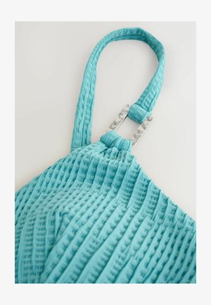 Sac en tissu turquoise texturé avec un motif unique. Présente un accent rectangulaire en argent au niveau de la jonction de la sangle pour un soutien.