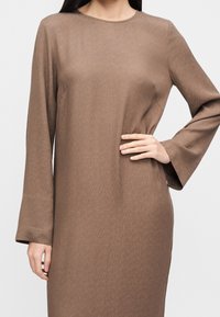 Femme portant une robe à manches longues, marron clair, texturée avec un col rond et une main posée sur sa hanche.