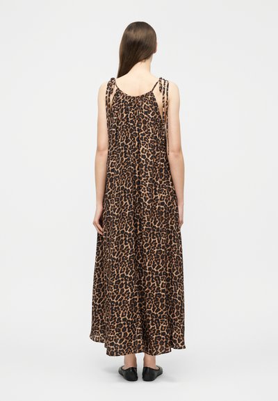 Γυναίκα στέκεται με την πλάτη γυρισμένη, φοράει μια χαλαρή, μακριά μέχρι τον αστράγαλο φορεματά με λεοπάρ σχέδιο, λεπτά κορδόνια στους ώμους και μαύρα παπούτσια χωρίς δέσιμο.