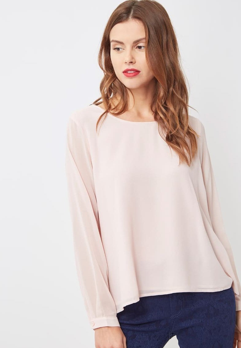 Mujer con cabello castaño ondulado, vistiendo una blusa de manga larga rosa claro y pantalones azul marino, mirando de lado frente a un fondo liso.