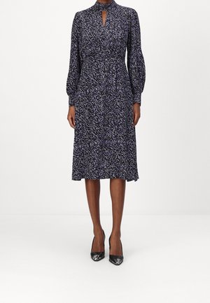 Vrouw draagt een knielange zwarte jurk met een paarse abstracte print, lange pofmouwen, een keyhole-halslijn en zwarte hoge hakken.