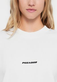 Biely sveter s okrúhlym výstrihom, s čiernym vyšitým logom "PEGADOR" v strede. Hladká bavlnená textúra, minimalistický dizajn.