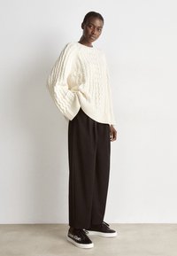 Krémový svetr s pleteným vzorem, volný střih, spárovaný s černými kalhotami s širokými nohavicemi a černými teniskami s bílým logem. Celkově hladký materiál.