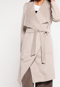 Cappotto avvolgente beige realizzato in tessuto morbido, con colletto drappeggiato, cintura legata in vita e due tasche frontali.
