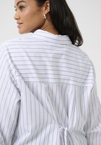 Camisa blanca de rayas con un detalle de ajuste en la parte de atrás, que presenta estrechas rayas verticales grises y un cuello. La tela parece ligera y texturizada.
