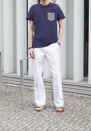 Personne debout à l'extérieur portant un t-shirt bleu marine avec une poche rayée, un pantalon blanc, des sandales et des bracelets devant une façade de bâtiment moderne.