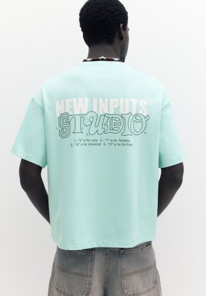 Homme portant un t-shirt bleu clair avec le texte « NEW INPUTS STUDIO » et la signification des lettres au dos, associé à un pantalon gris et un collier de perles.