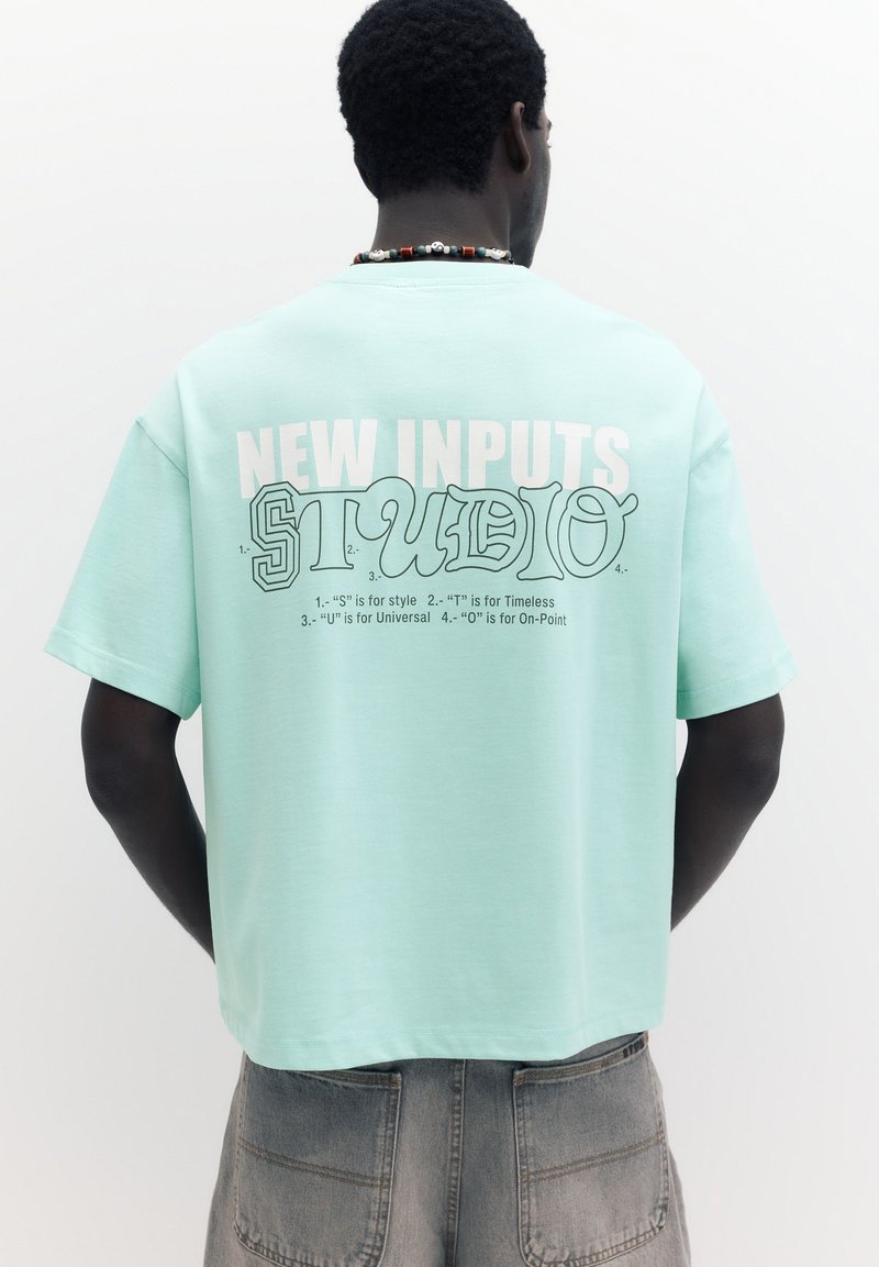 Homme portant un t-shirt bleu clair avec le texte « NEW INPUTS STUDIO » et la signification des lettres au dos, associé à un pantalon gris et un collier de perles.