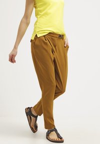 Person med senapsgula plisserade byxor, gul kortärmad topp och svarta sandaletter med öppen tå går mot en vit bakgrund.