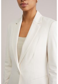 Inimese ülisuuremõõtmeline pilt, kes kannab kreemika värvi bleiserit beežikat ribilise mustriga särgi peal, rõhutades bleiseri krae ja ühe nupuga sulgemist.