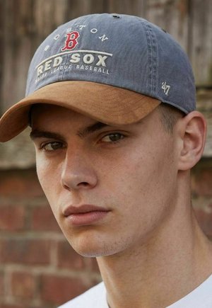Junger Mann mit einer blau-braunen Boston Red Sox Baseballkappe, neutralem Gesichtsausdruck, vor einem verschwommenen Ziegelwand-Hintergrund.